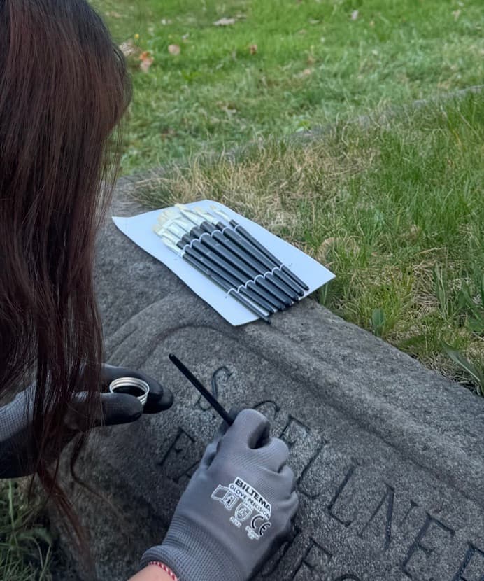 Minnesfrid fyller i bokstäver på en gravsten med tunn pensel och skyddshandskar